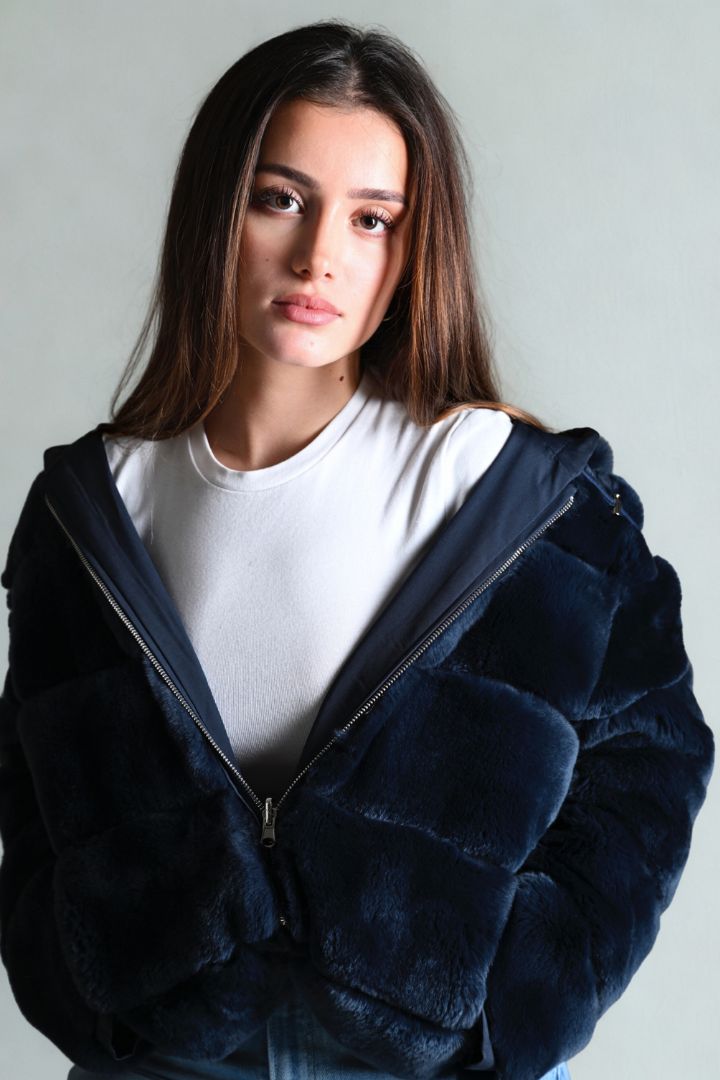 Woman wearing a dark blue fur coat over a white shirt against a plain background
