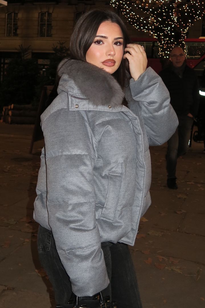 Woman wearing a grey down puffer jacket with a fox fur collar, posing outdoors at night with festive lights in the background.