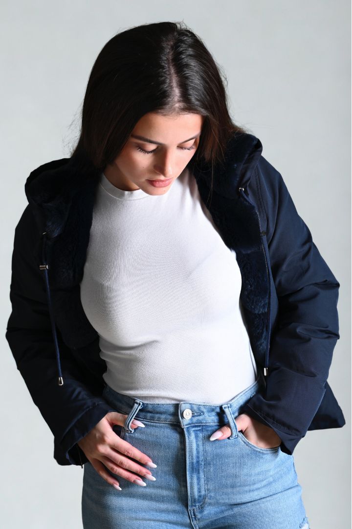 Woman wearing a navy jacket, white shirt, and blue jeans against a plain background