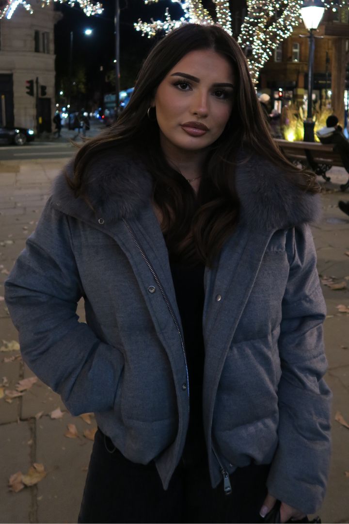 Front view of a woman wearing a grey fox fur–collared puffer jacket, standing in a city street illuminated by warm night lights.