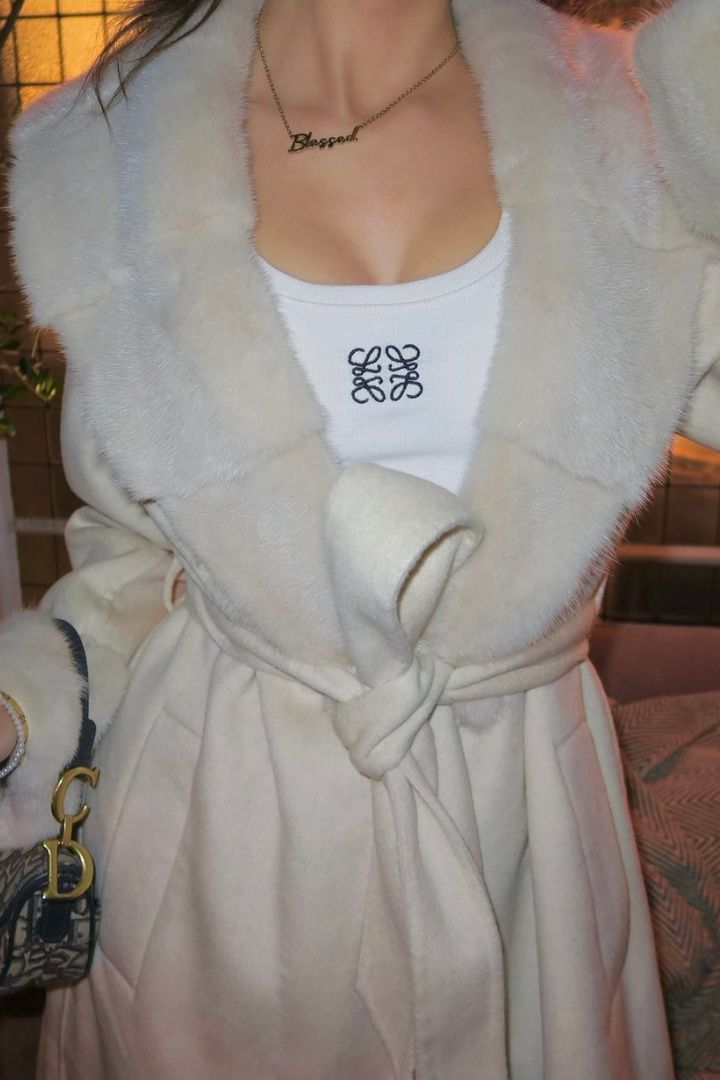Close-up of beige faux-fur trim coat tied at the waist with a white top underneath and designer bag detail.