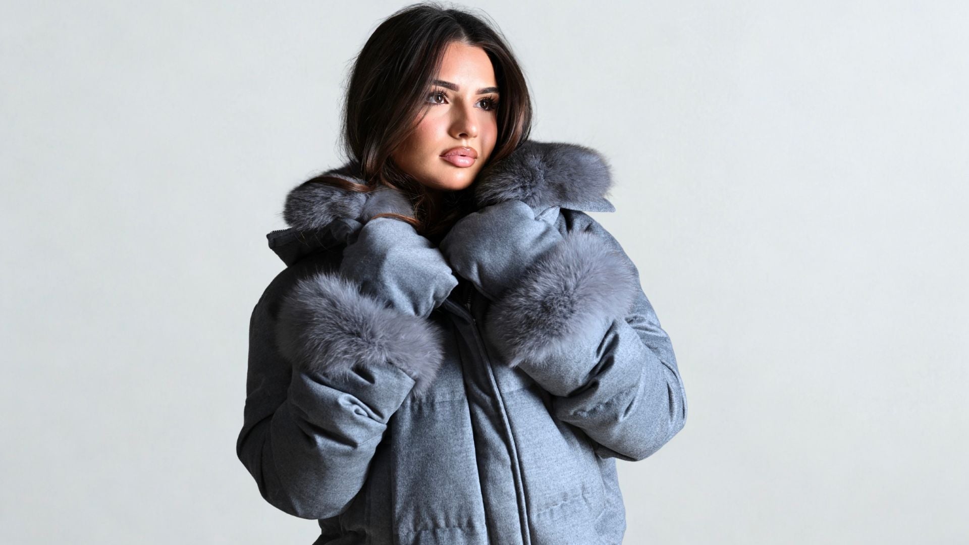 A woman wearing a grey winter coat with plush faux fur trim on the hood and sleeves, looking off to the side while holding the collar for warmth.
